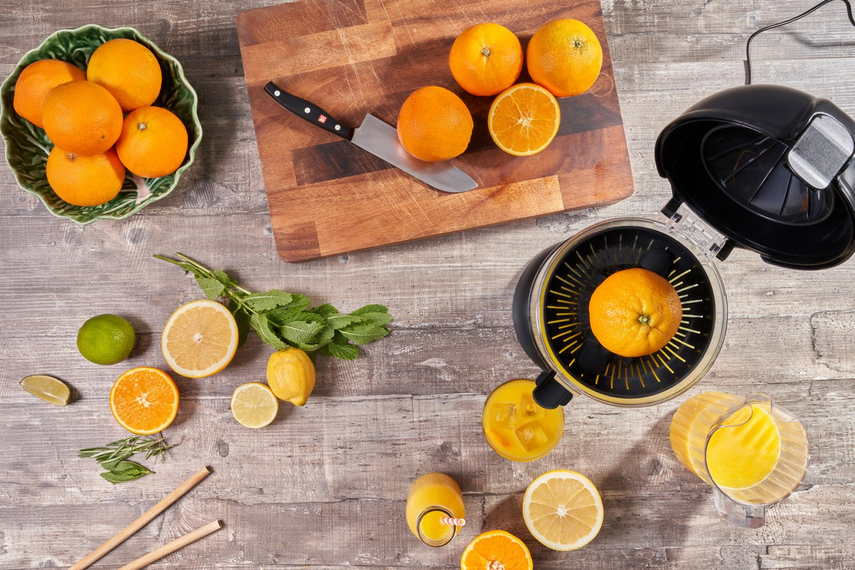 Fridja f900 automatic citrus juicer with a halved orange, surrounded by oranges, lemons, limes, fresh mint and glasses of juice on a wooden table