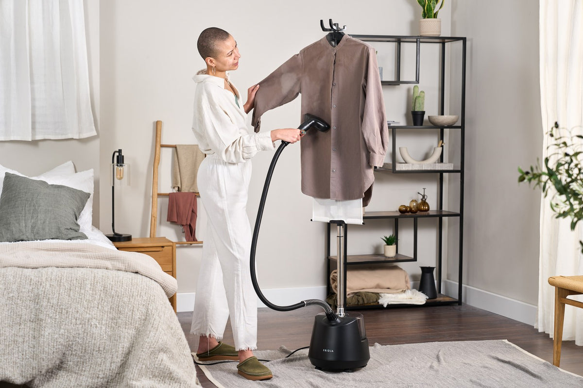 Woman steaming a long taupe shirt on the hanger of the Fridja f1200 upright clothes steamer in a bright bedroom