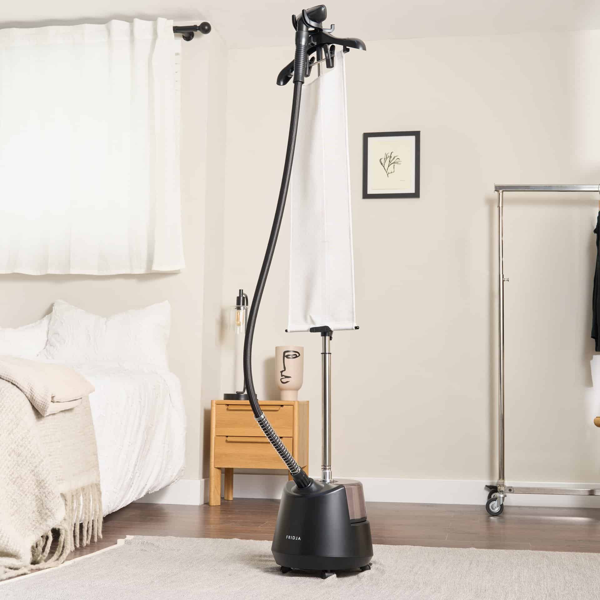 A refurbished Fridja f1200 Clothes Steamer Pro stands in a neat bedroom by the bed, nightstand, and clothes rack. Sunlight filters through white curtains, and minimalist art adds style to this premium steaming setup.