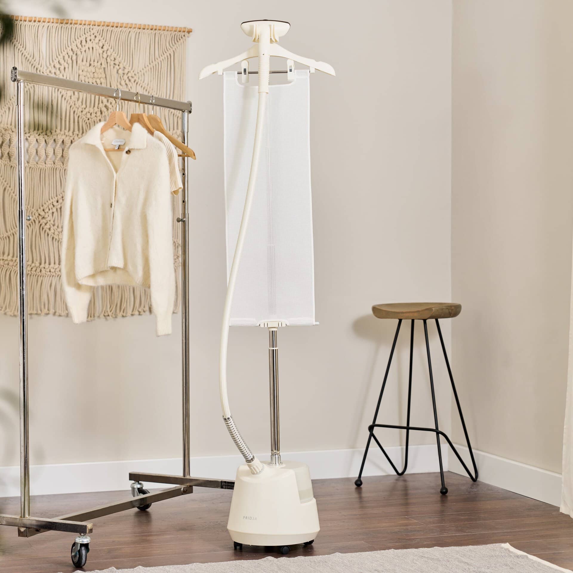 The Fridja f1200 Clothes Steamer Pro - Refurbished stands next to a clothing rack with sweaters, a wall hanging, and a wooden stool on a hardwood floor with a rug, providing a premium steaming experience.