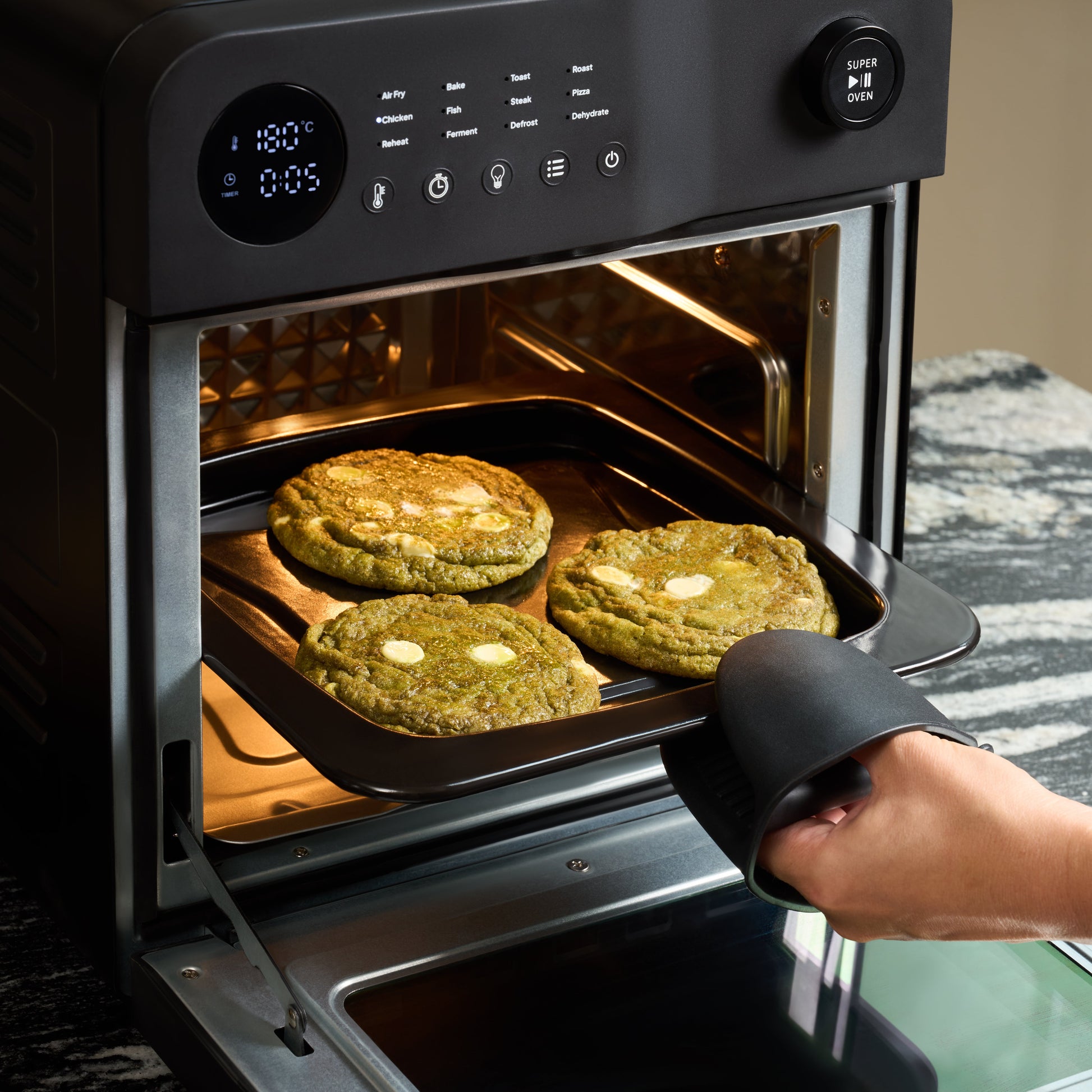 A person wearing a black oven mitt pulls out a tray with three large green cookies from a Fridja f66 Super Oven - 15L Digital Air Fryer Oven, whose display shows 18:05.