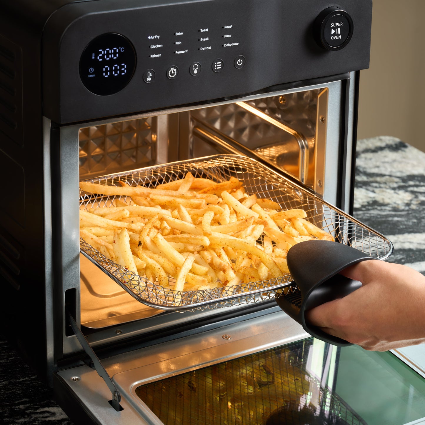A hand in an oven mitt removes cooked fries from a Fridja f66 Super Oven - 15L Digital Air Fryer Oven, which features a PFAS free design and a digital display showing temperature and time for precise convection cooking.