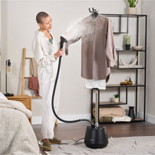 A person uses the Fridja f1200 Clothes Steamer Pro - Refurbished Grade A to remove wrinkles from a brown shirt on a steamer stand in a modern bedroom, highlighting professional-grade garment care with visible steam.