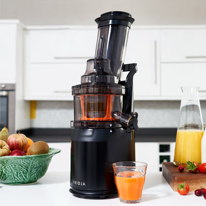 The Fridja Juicer Duo – f1900 Cold Press Juicer & f500 Portable Blender sits on a kitchen counter, pouring fresh orange juice into a glass with mixed fruit, a carafe of juice, and strawberries nearby. White cabinets are in the background.