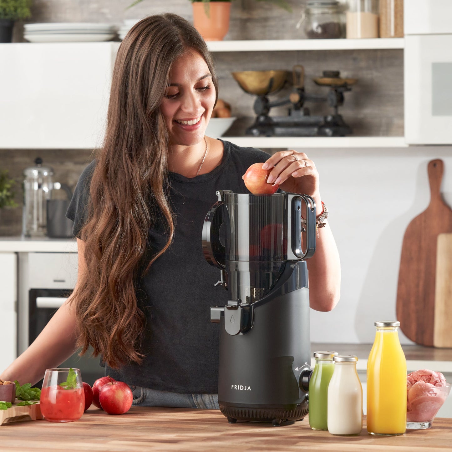 A woman with long brown hair smiles as she places an apple into a Fridja f2500 Self Feeding Whole Fruit Juicer - Refurbished in a modern kitchen, with fresh apples, leafy greens, and cold-pressed juice on the wooden counter nearby.