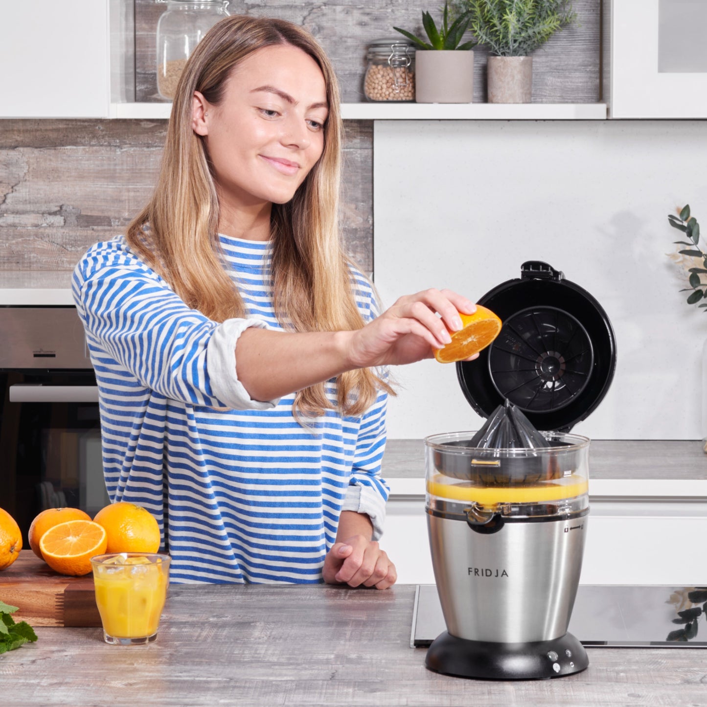 A woman in a striped shirt uses the Fridja f900 Automatic Citrus Juicer - Refurbished to squeeze an orange in a modern kitchen, with a glass of juice and whole oranges on the counter beside her.